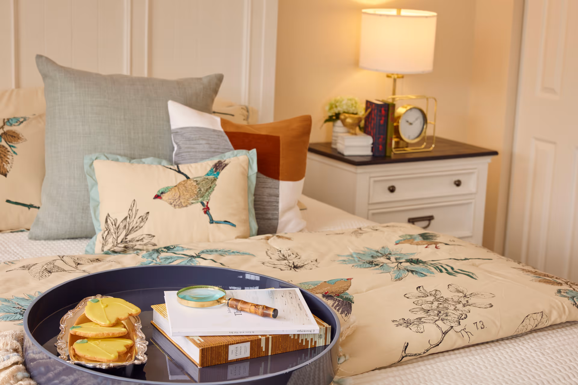 A cozy bedroom scene featuring a bed with multiple decorative pillows, including one with a bird design, and a floral-patterned comforter. On the bed is a round tray holding a few books, a magnifying glass, and a plate with yellow-frosted cookies. In the background, there is a white nightstand with a lamp, a clock, some books, and a small plant.