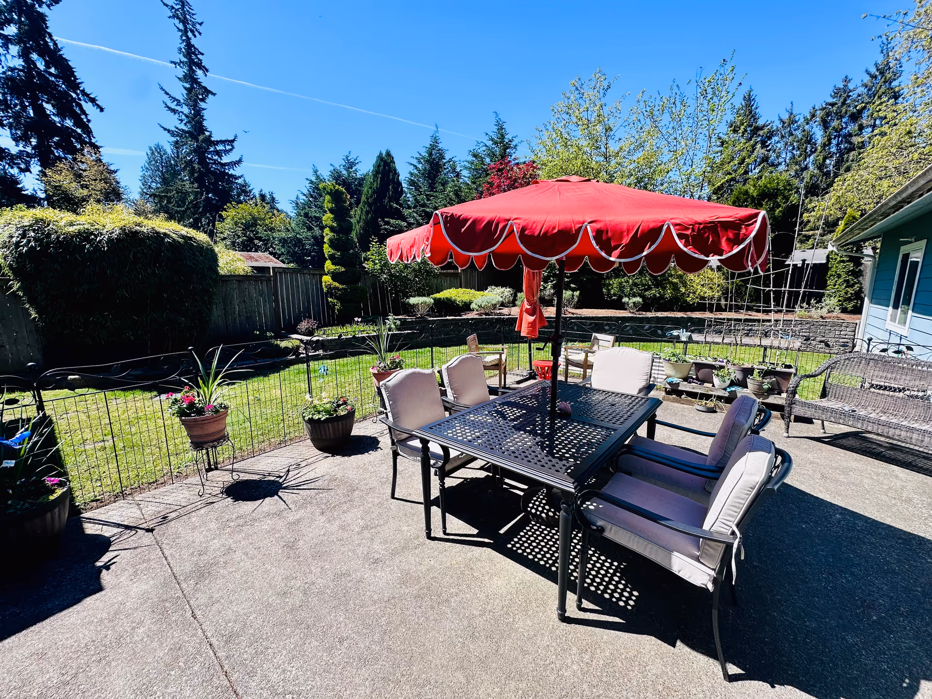 Outdoor patio with red umbrella, metal table, cushioned chairs, potted plants, and a green yard.