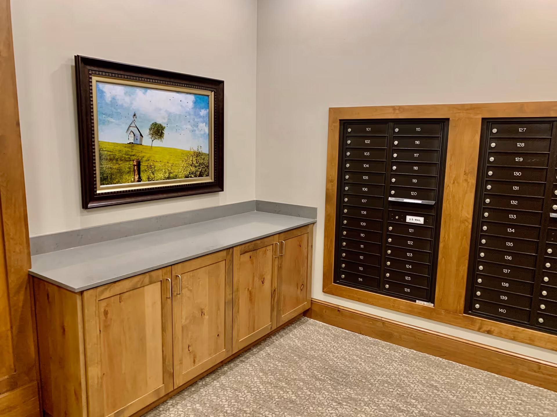 Interior corner of a facility with a wooden cabinet topped with a gray countertop, a framed picture of a small white church and tree on a grassy hill hanging above it, and a wall-mounted set of black mailboxes with numbered slots.