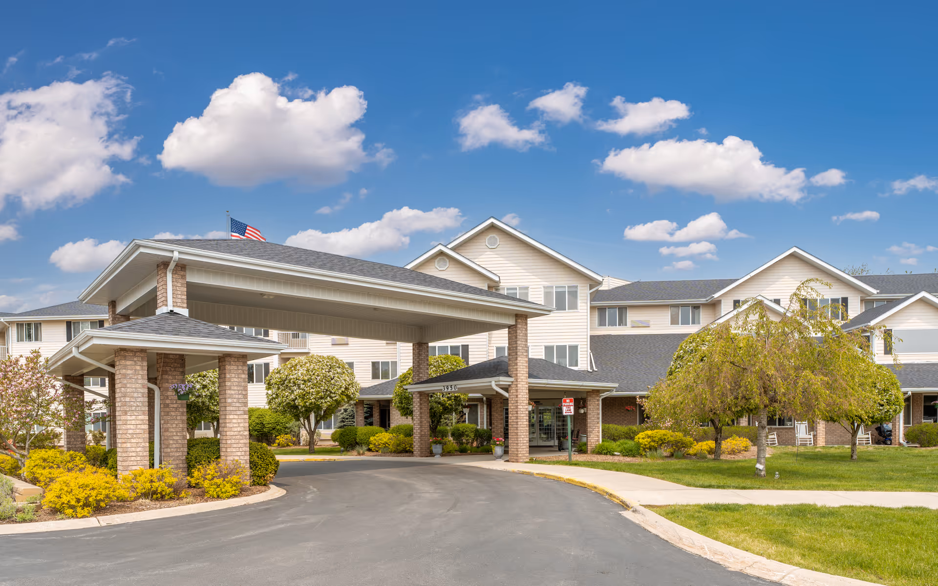 Covered entrance and driveway of a multi-story senior living building with landscaped grounds and an American flag.