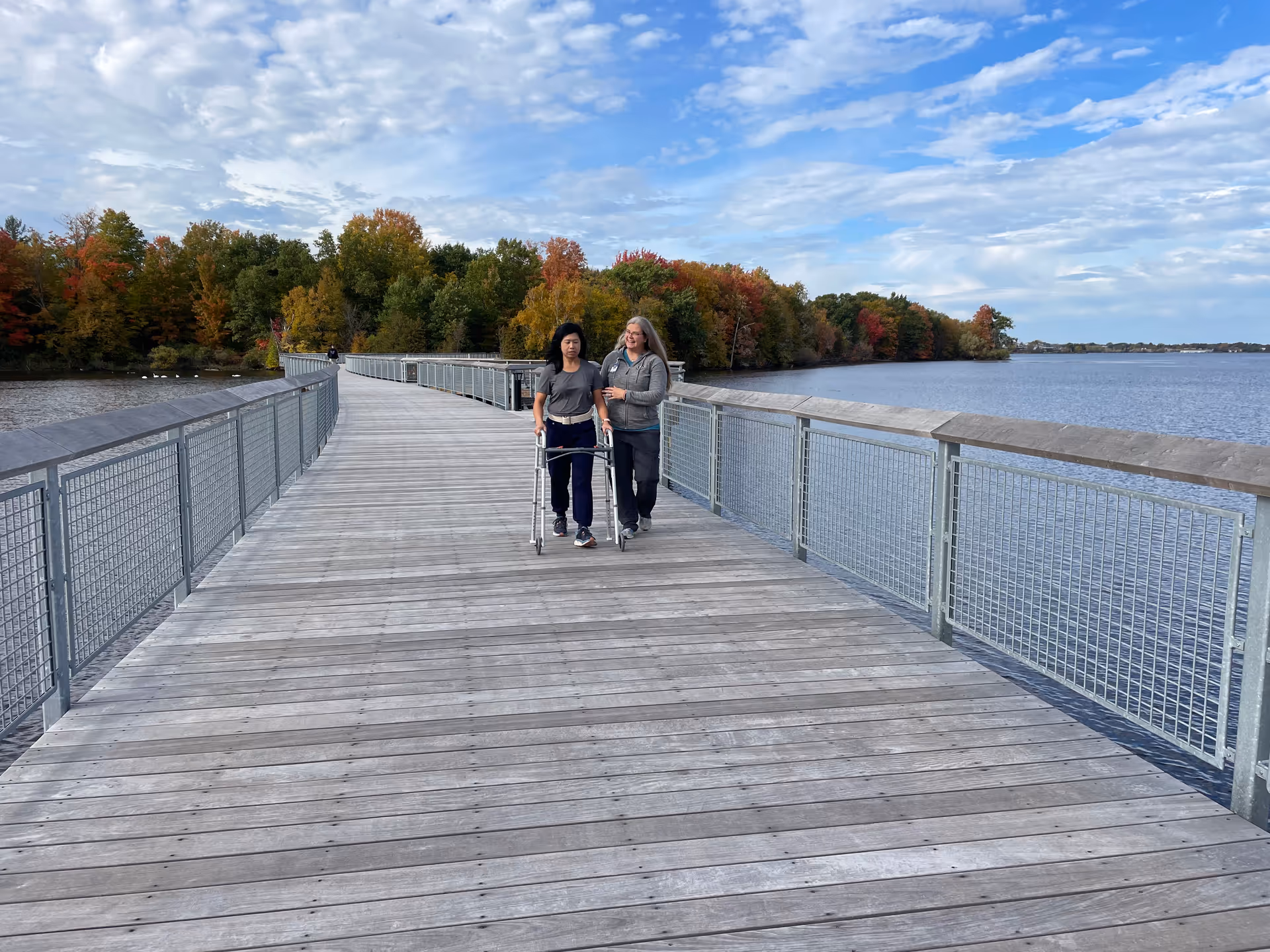 Two people walking with a walker on a wooden boardwalk by a lake with autumn trees.