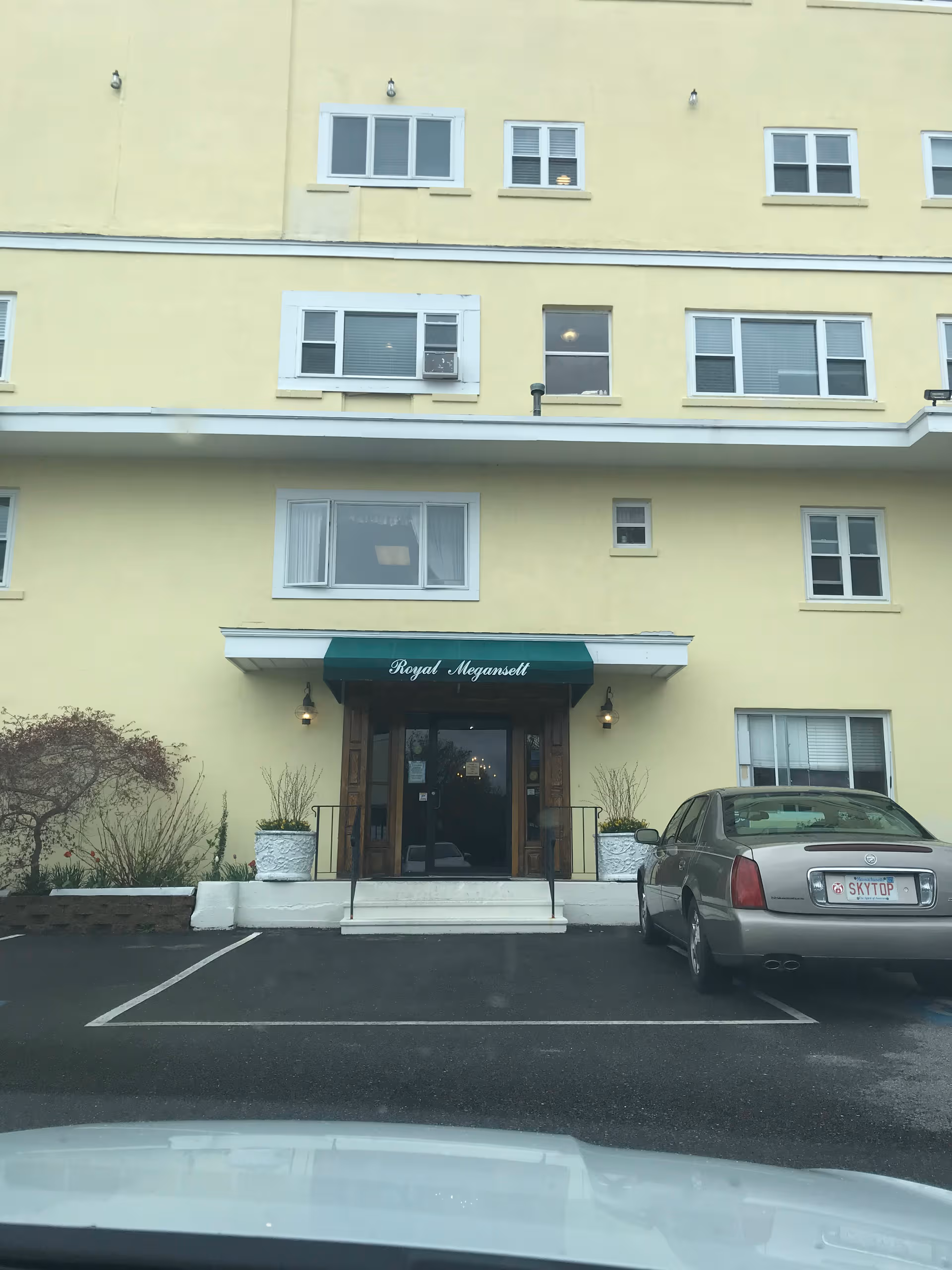 Yellow multi-story building with entrance, green awning reading Royal Megansett, parking lot in front.