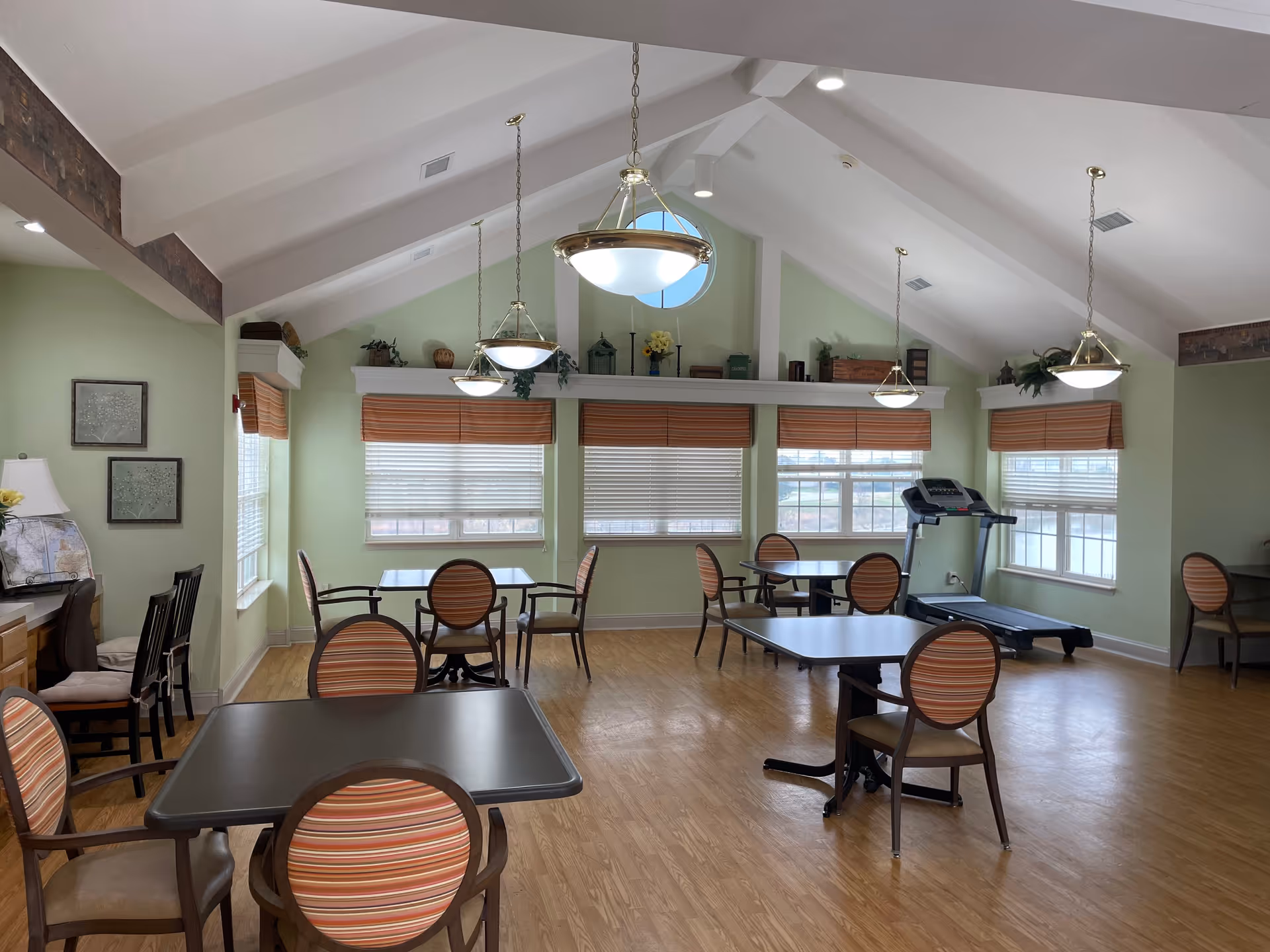 Bright senior living common area with round tables, striped chairs, blinds, and a treadmill.