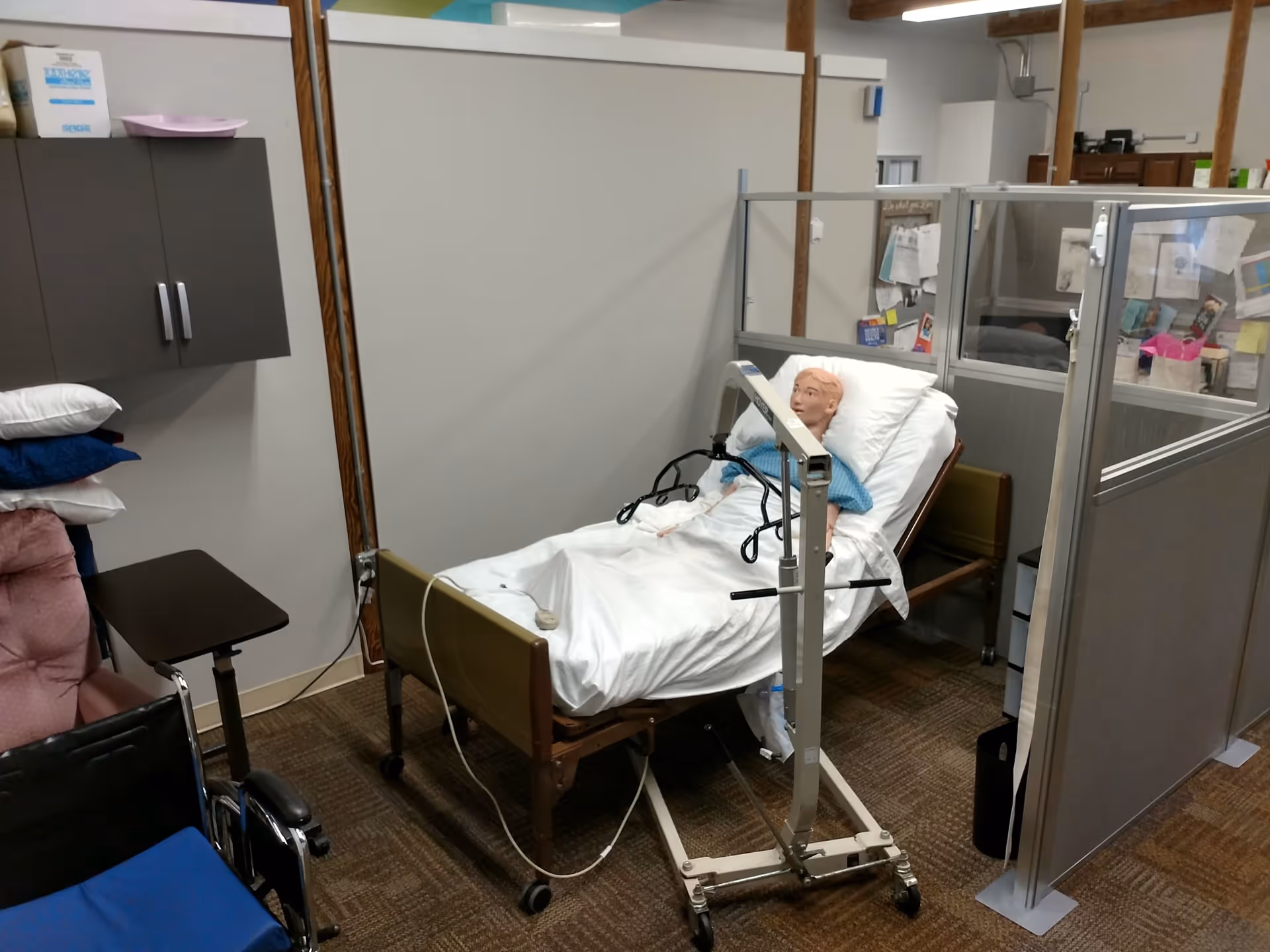 Medical training mannequin lies in a bed within a partitioned, cubicle-style care room.