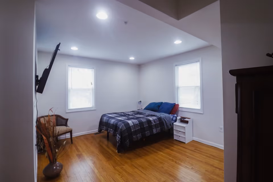 Bedroom in a senior living facility with a bed, nightstand, dresser, and mounted TV.