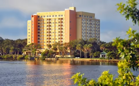 Tall pastel-colored senior living building beside a lake with palm trees.