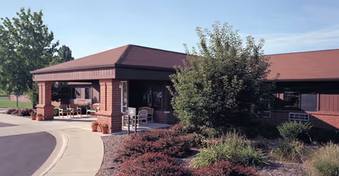 Front entrance of a single-story brick senior living facility with a covered drive-up area, benches, and landscaped shrubs.