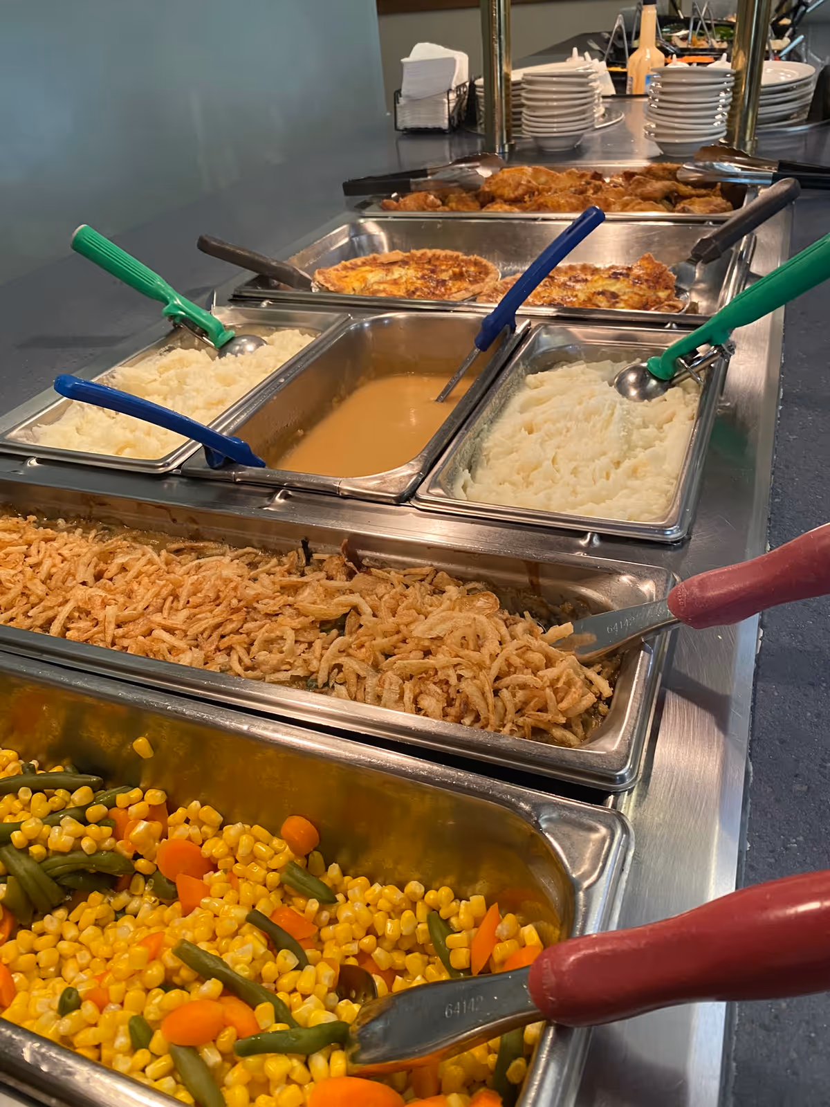 Buffet-style serving line with trays of mixed vegetables, fried onions, mashed potatoes, gravy and other hot dishes.