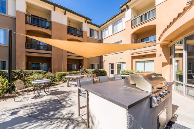 Sunny courtyard patio with a built-in grill, tables and chairs, a large shade sail, and surrounding three-story residential building.