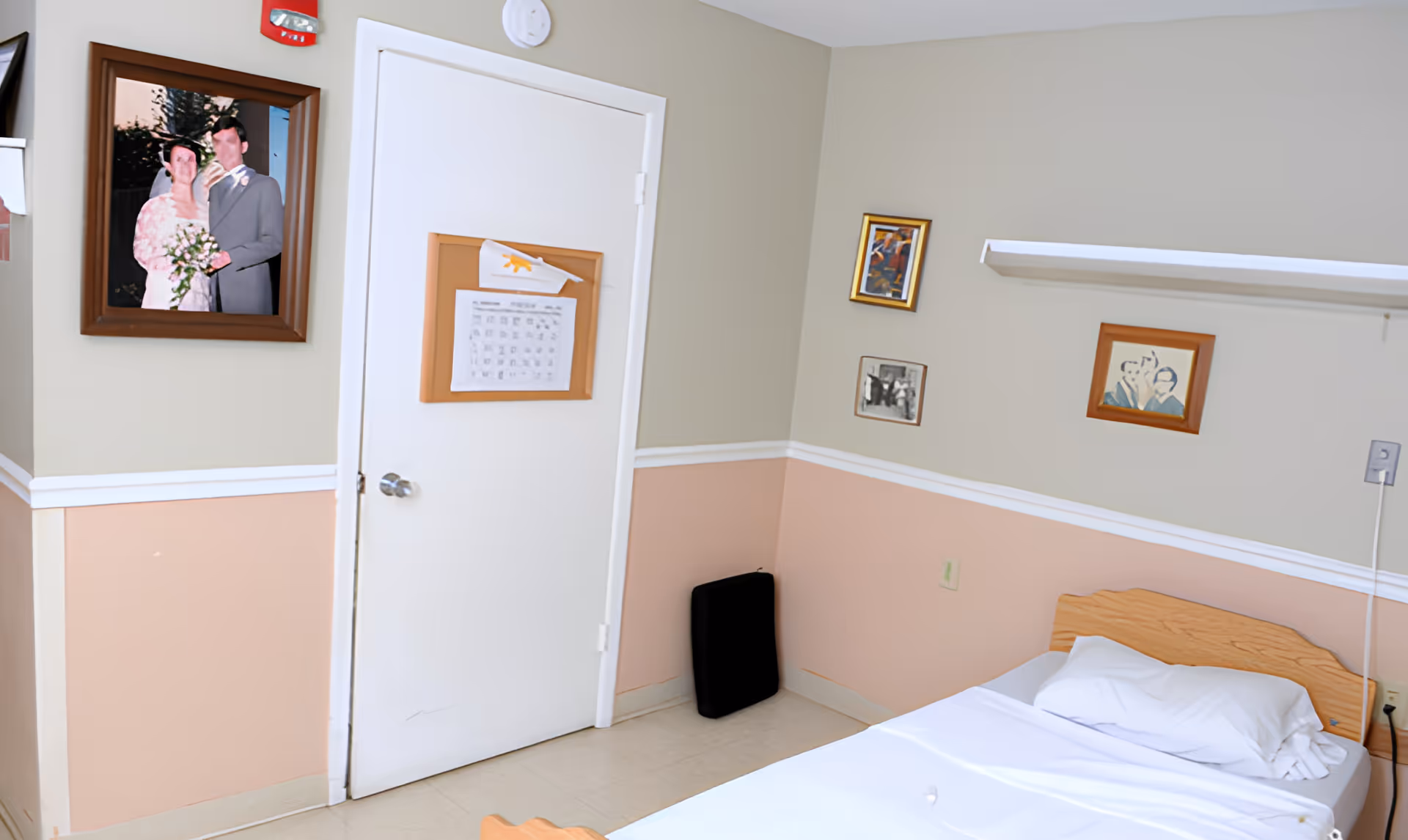 A simple nursing home bedroom with a single bed made with white sheets and a pillow. The walls are painted in two tones, beige on top and peach on the bottom, separated by a white chair rail. There are several framed pictures hanging on the walls, including a wedding photo and some artwork. A white door has a corkboard with papers pinned to it. A black cushion or pad rests against the corner of the wall near the door.