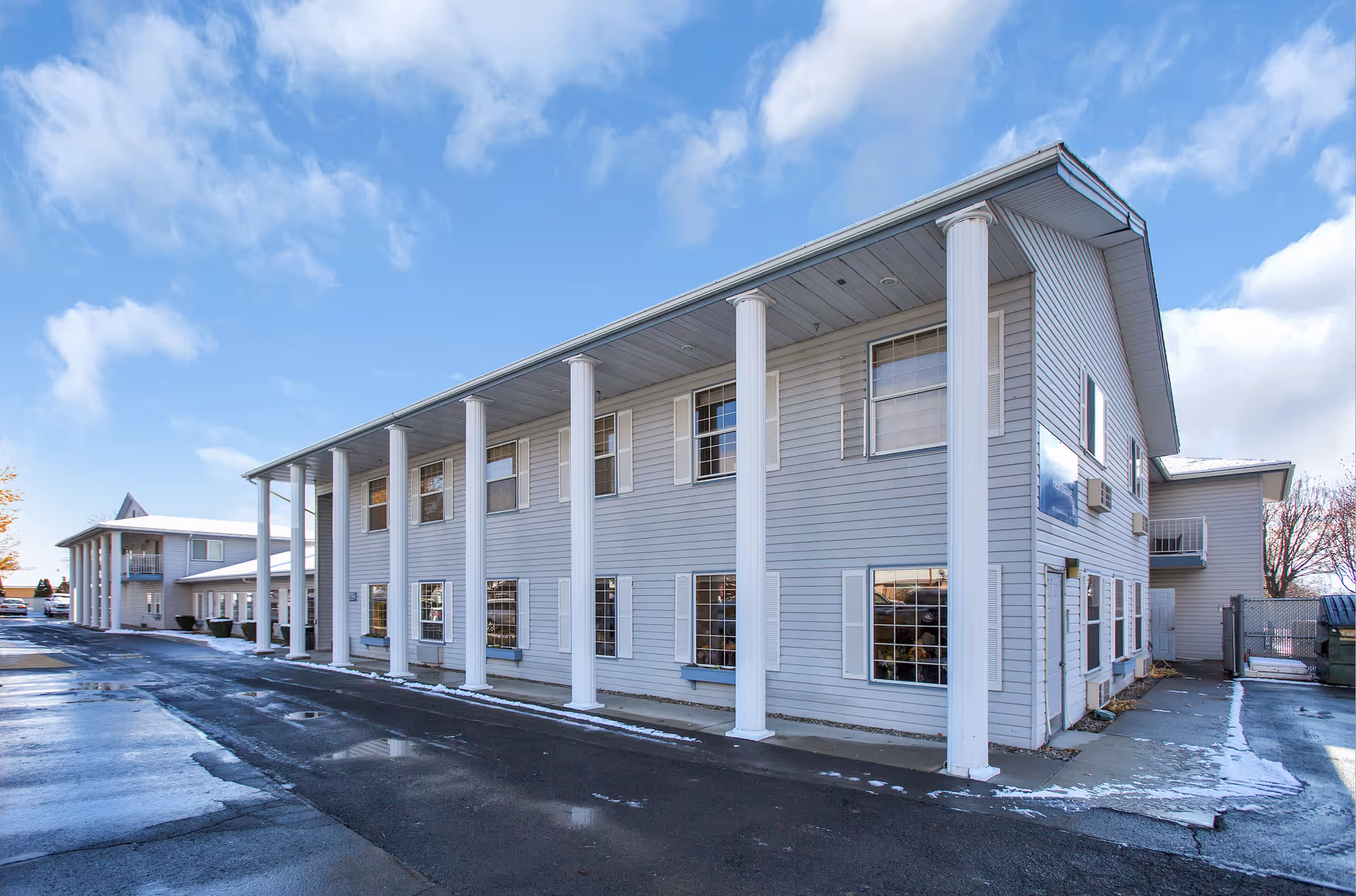 Exterior view of a two-story senior living facility building with white siding and tall white columns along the front. The sky is partly cloudy and the ground shows patches of snow and wet pavement.