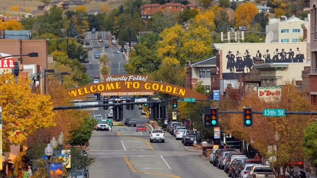 A street view of downtown Golden with a large archway sign reading 'Howdy Folks! Welcome to Golden Where the West Lives'. The street is lined with parked cars and trees with autumn foliage. Buildings and hills are visible in the background.