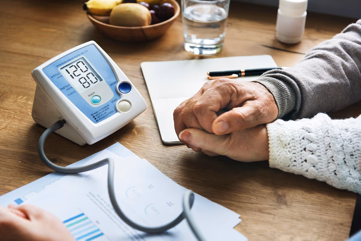 Elderly hands resting near a digital blood pressure monitor on a wooden table.