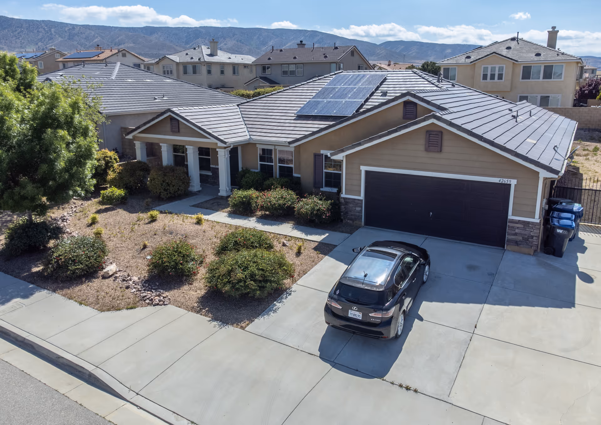 Front exterior of a single-story suburban house with solar panels on the roof, a two-car garage, a car parked in the driveway, and desert landscaping.