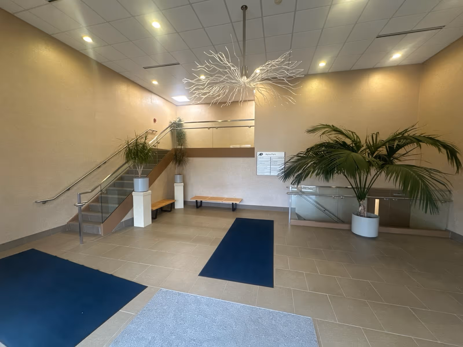 Bright, spacious lobby with stairs, glass railing, benches, and potted palm plants.
