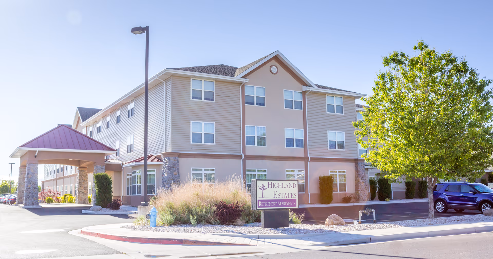 Exterior view of a three-story retirement apartment building named Highland Estates with a covered entrance, parking area, landscaping including a tree and bushes, and a clear blue sky.