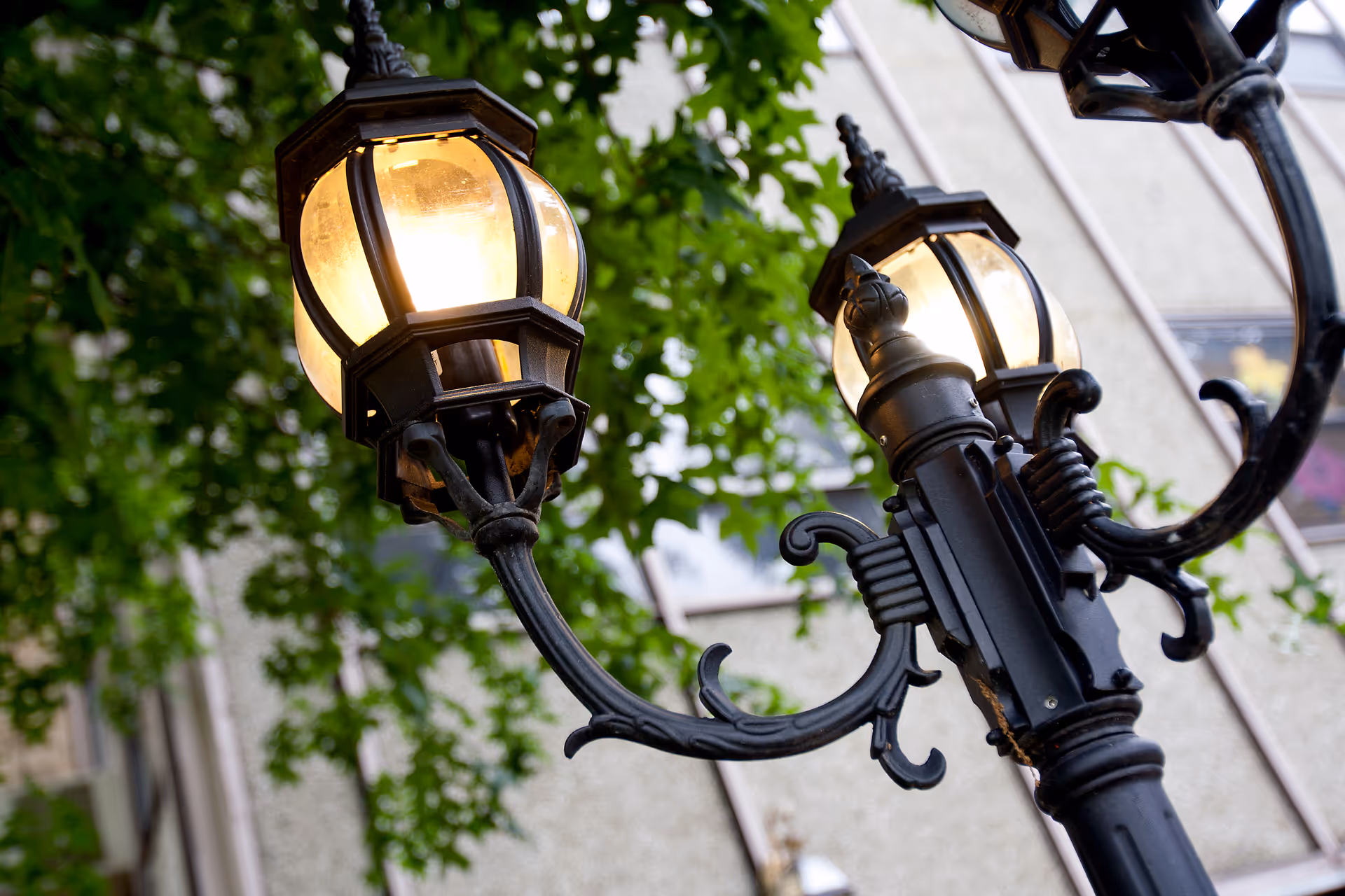 Ornate street lamp with glowing bulbs against a building and greenery