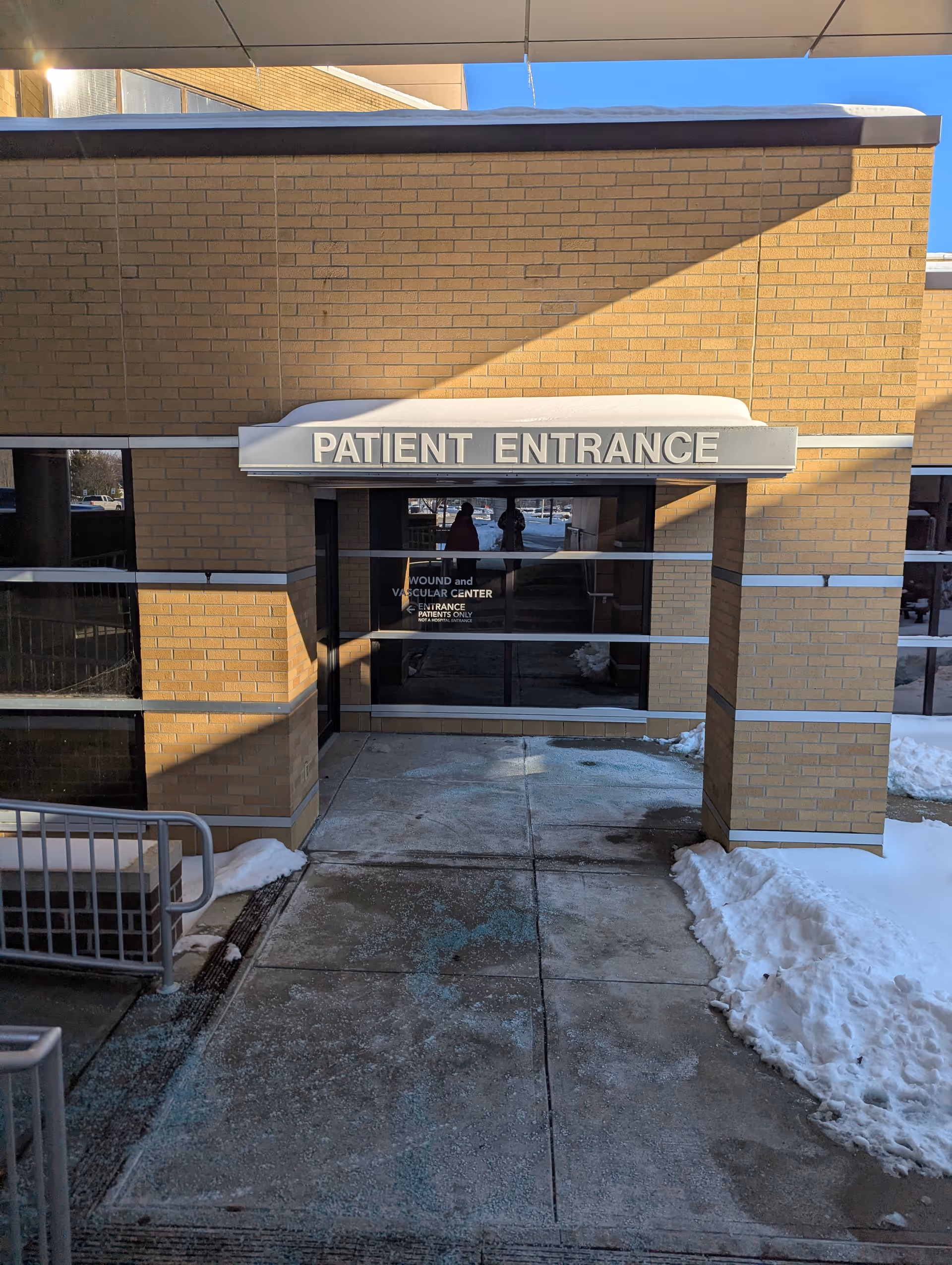 Exterior brick building entrance with a 'PATIENT ENTRANCE' sign and snow on the sidewalk.
