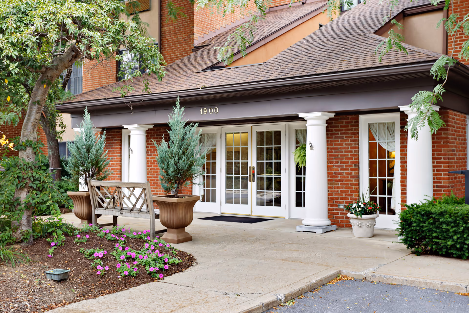 Entrance of a senior living facility with a brick exterior, white columns, and double glass doors. There are potted plants, a wooden bench, and a small garden with pink flowers near the walkway.