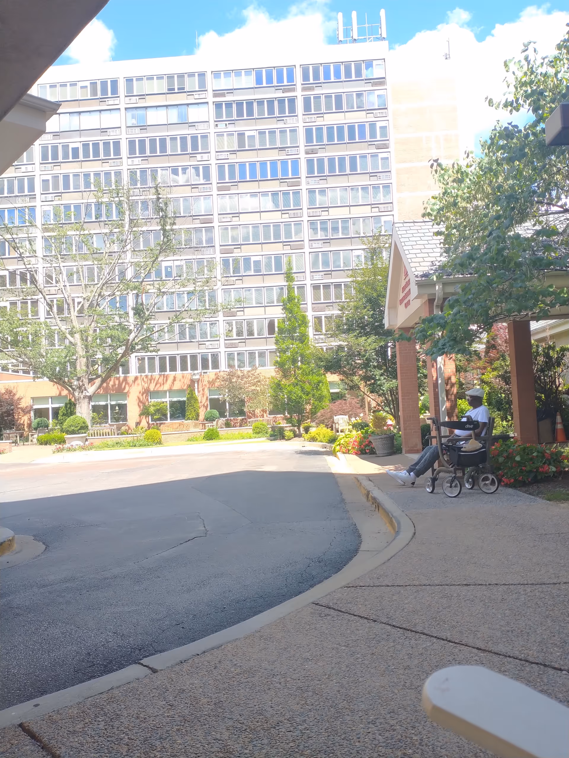 Exterior view of a senior living campus with a wheelchair-bound resident at a covered outdoor area.
