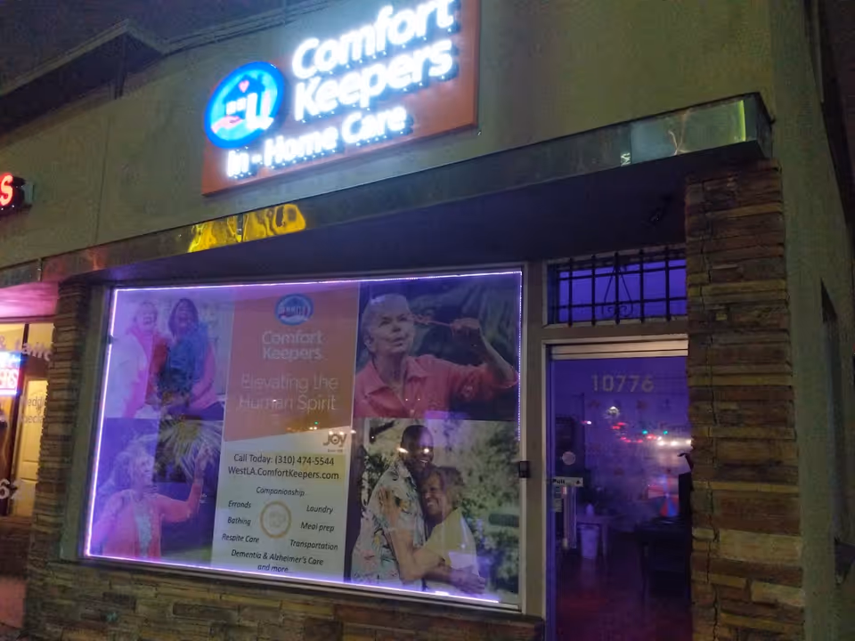 Night exterior of Comfort Keepers storefront with neon-lit signage and large window advertisement.
