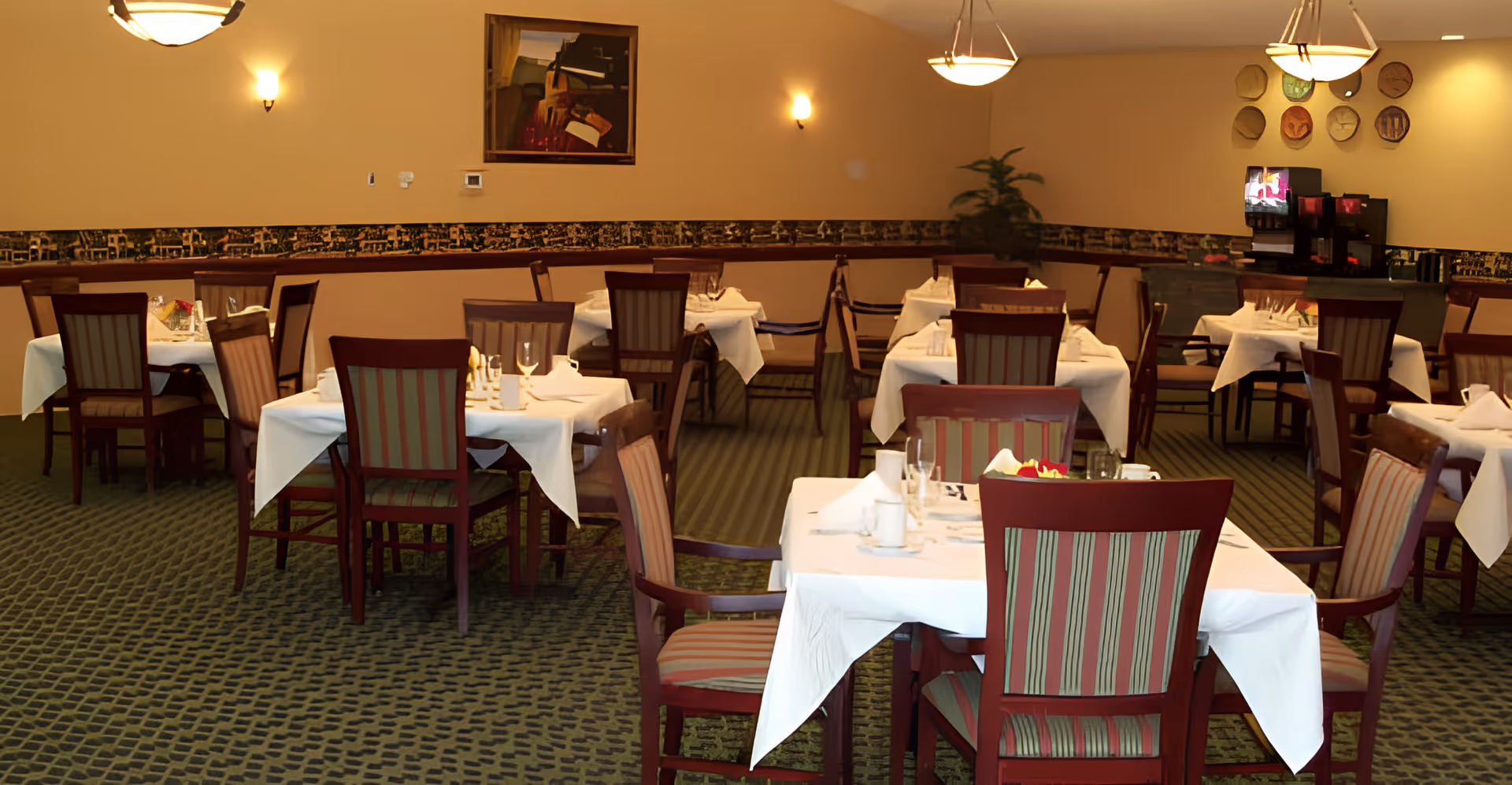 Dining room with multiple tables covered in white tablecloths, set with glassware and napkins. Wooden chairs with striped upholstery surround the tables. The room has warm lighting from ceiling fixtures and wall sconces, a patterned carpet, and decorative wall art including a framed painting and a collection of plates.