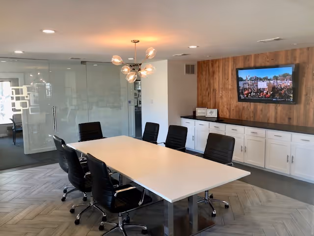 A modern meeting room with a long white table, black chairs, and a wall-mounted TV.