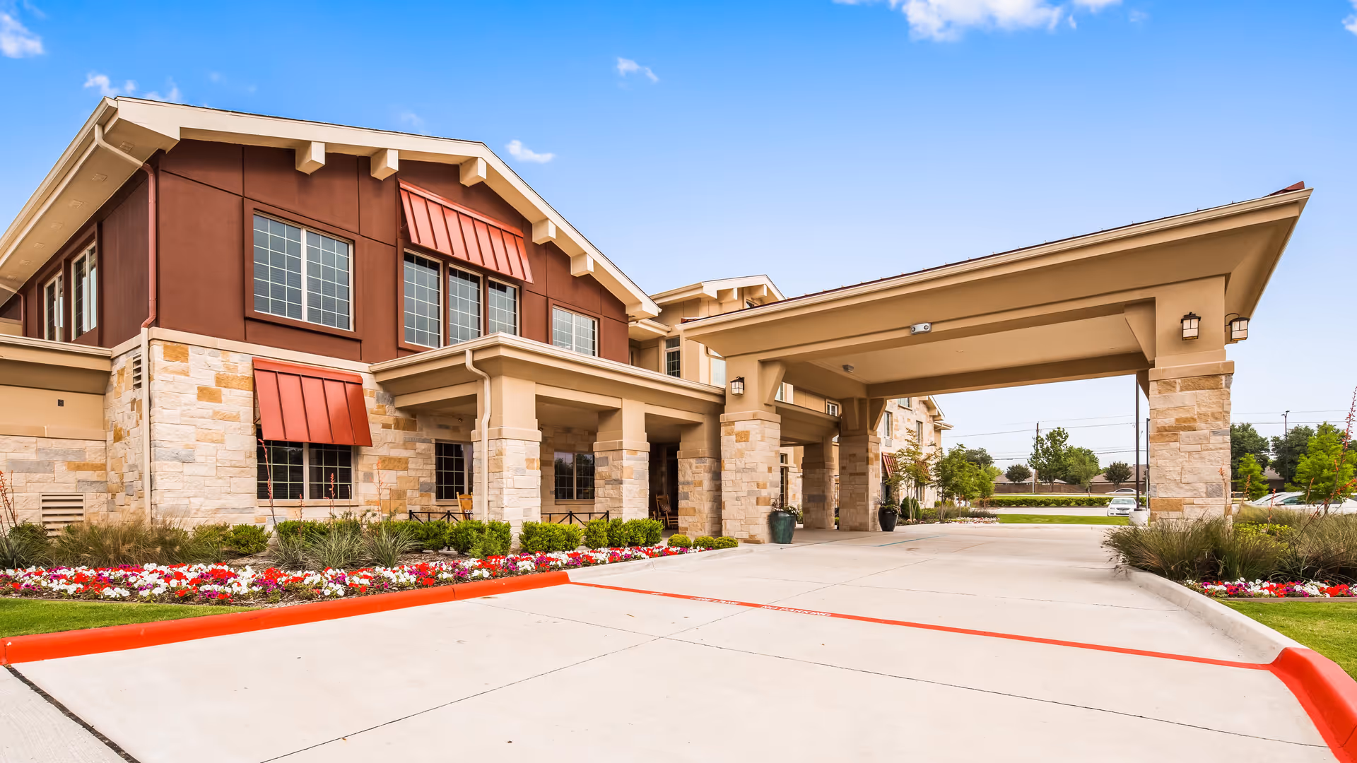 Exterior view of a senior living facility building with stone and brown siding, red window awnings, a covered entrance with pillars, and landscaped flower beds along the driveway under a blue sky.