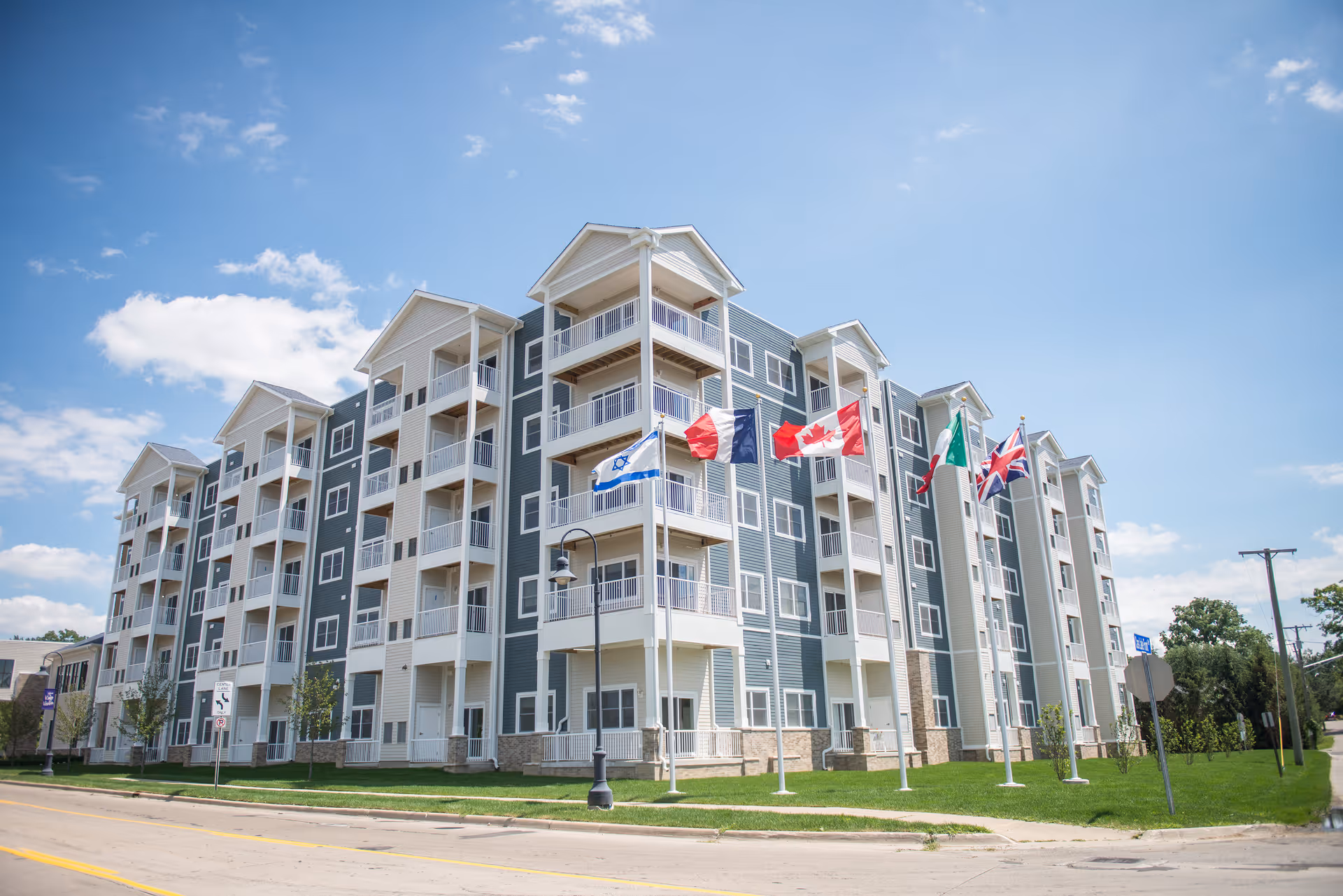 Exterior view of a five-story apartment building with balconies and several flagpoles in front under a blue sky.