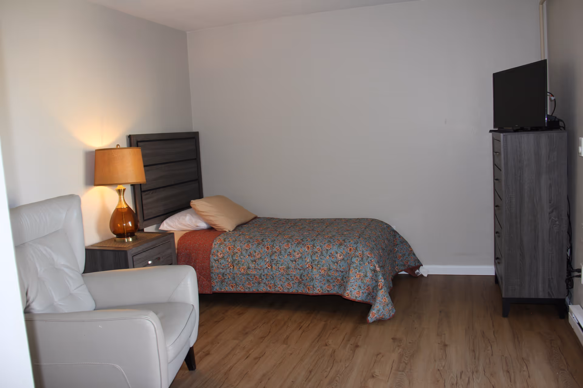 Bedroom with bed, nightstand lamp, dresser with TV, and white armchair.