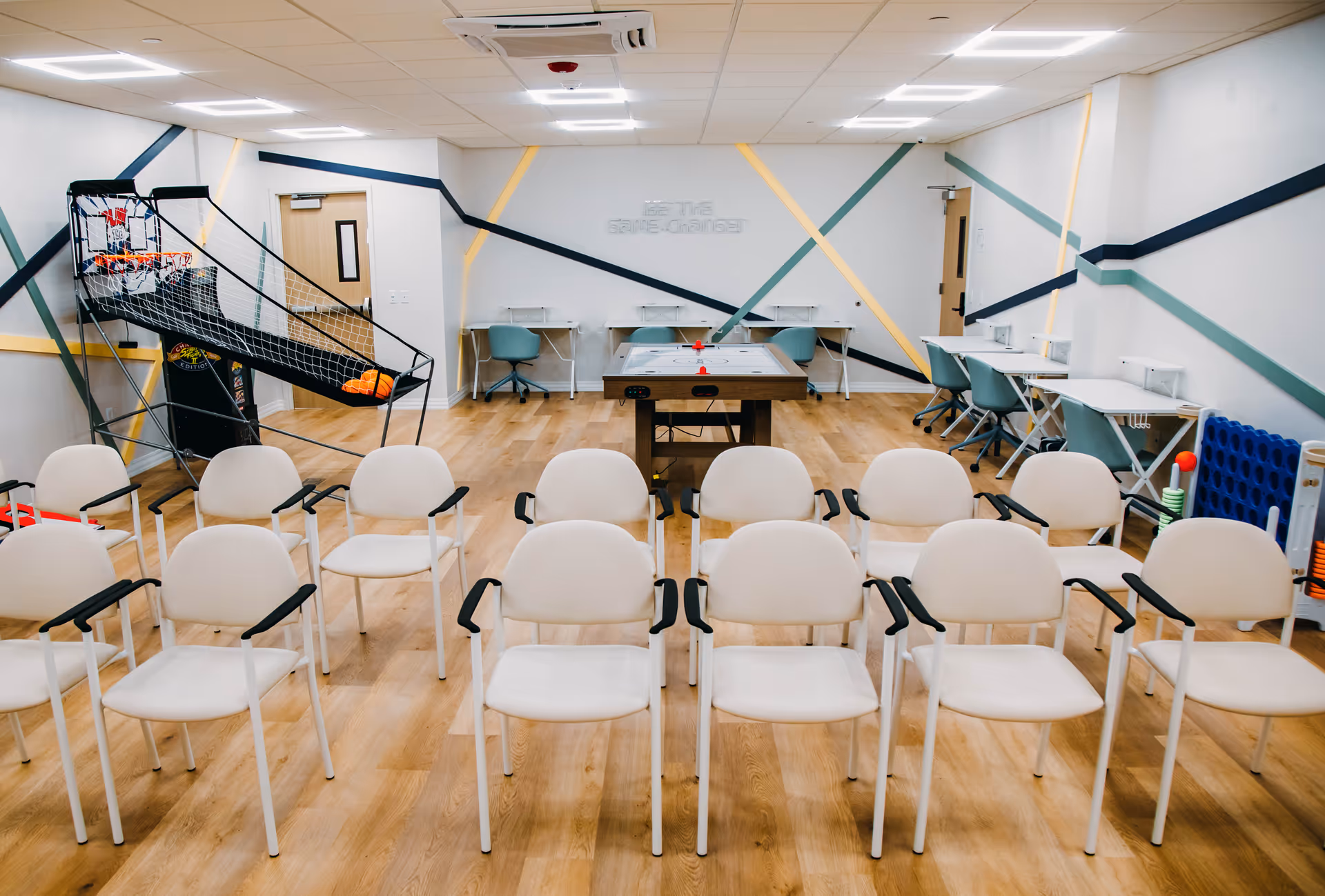 Empty recreation room with rows of white chairs, an arcade basketball game, and a pool table.