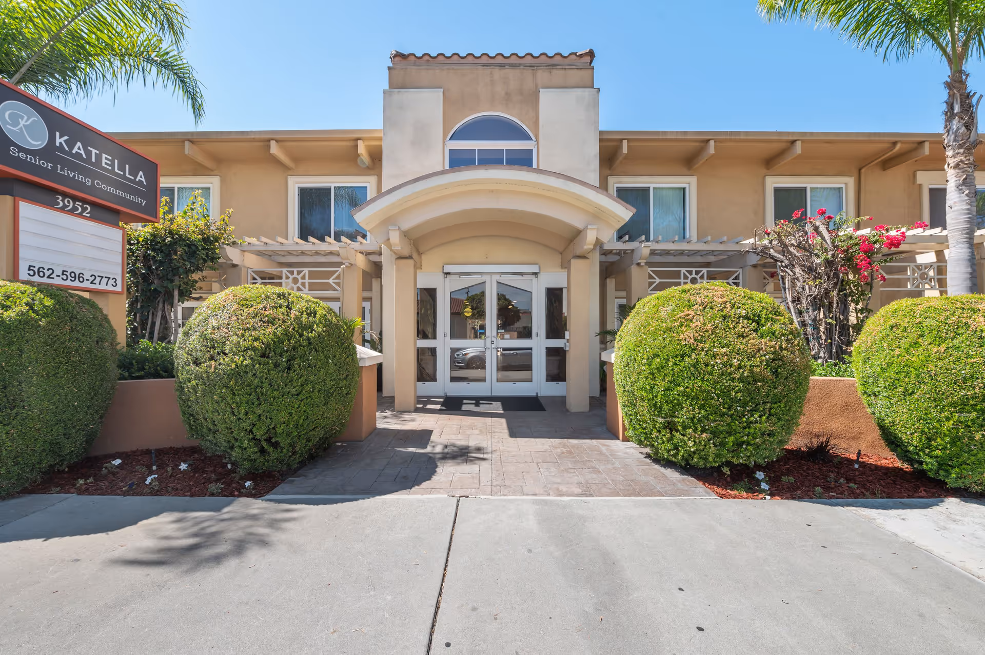 Front entrance of a beige senior living building with double glass doors, trimmed spherical shrubs, and a sign reading "Katella Senior Living Community".