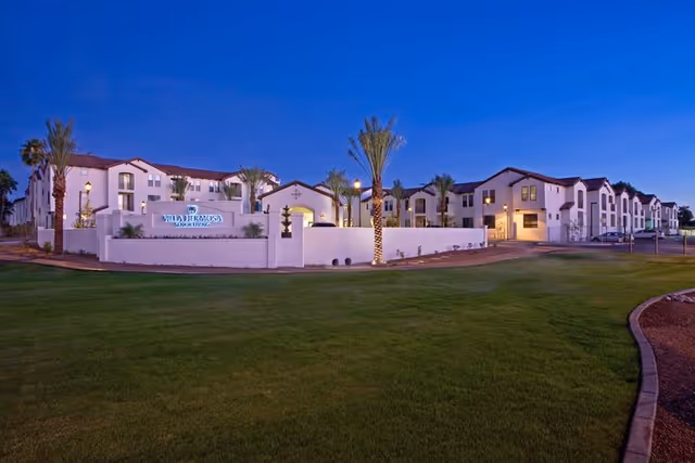 Exterior view of Villa Hermosa senior living facility at dusk, showing a large white building with multiple windows, palm trees, well-maintained green lawn, and a sign with the facility name at the entrance.