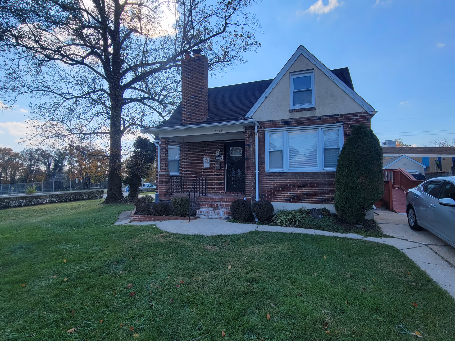 Brick cottage-style building with a manicured lawn, large tree, and driveway with parked car.