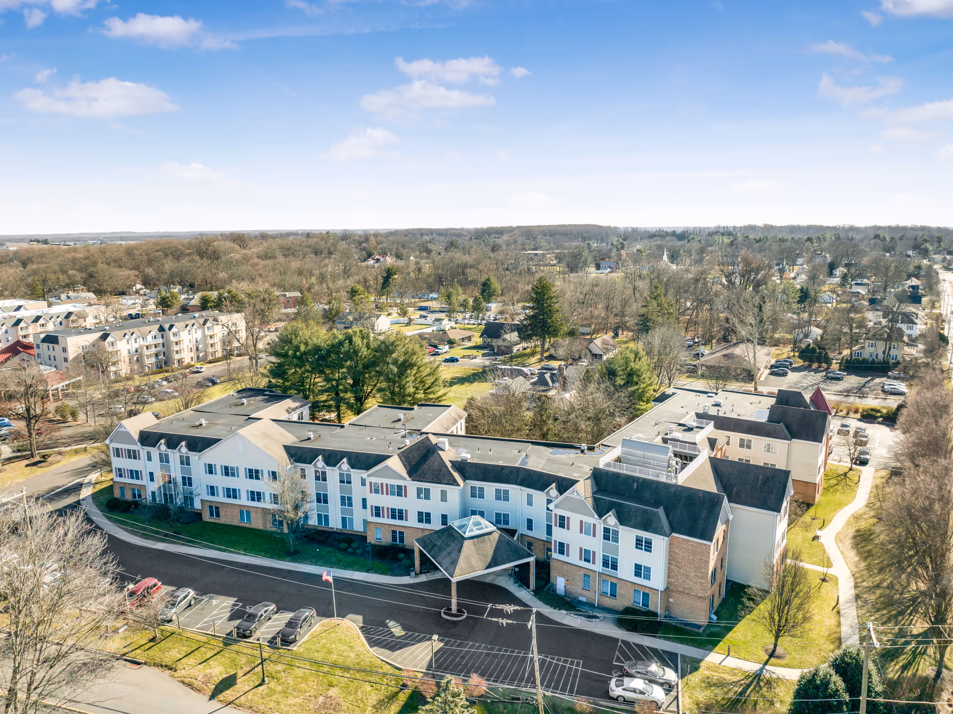 Aerial view of a large senior living facility named Oxford Crossings, showing a multi-story building with a covered entrance, surrounded by parking lots, trees, and residential neighborhoods under a partly cloudy sky.