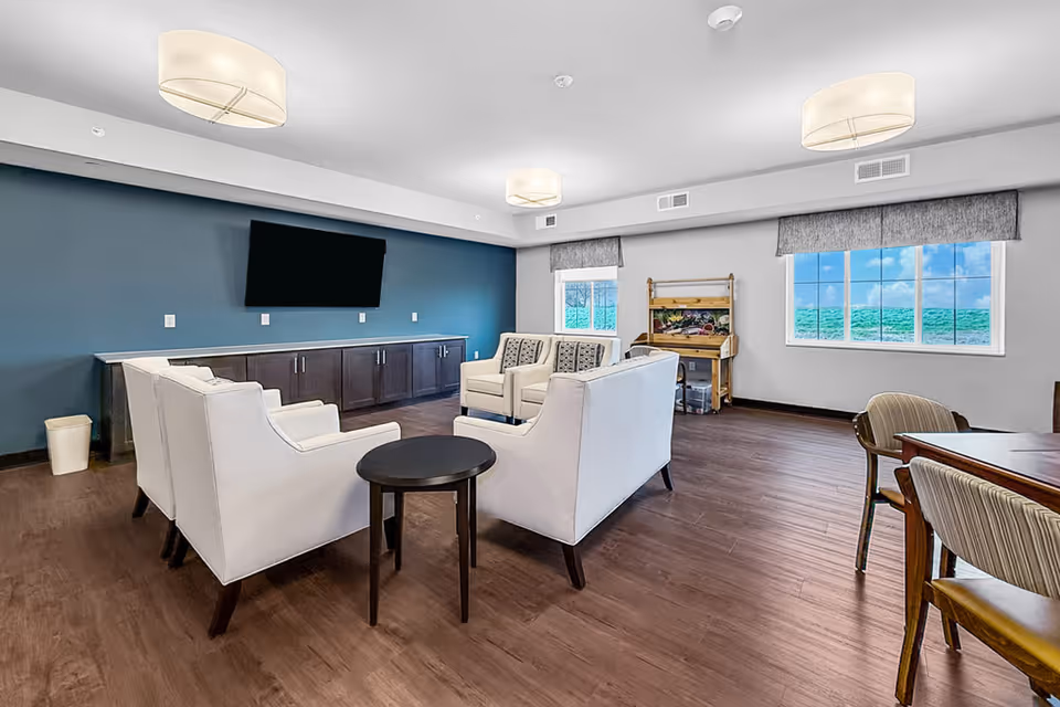 Senior living lounge with white sofas, teal accent wall, TV, and windows.