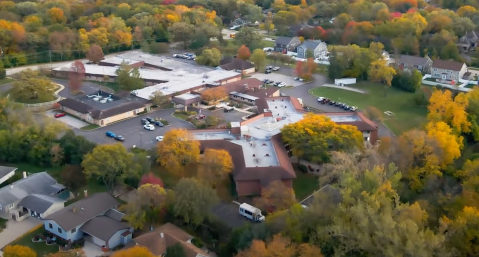 Aerial view of a senior living campus with connected buildings, parking lots, and fall foliage surrounded by houses.