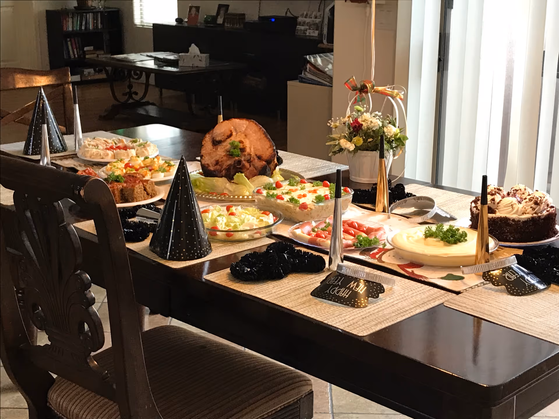 A dining table set with various dishes including a ham, salad, deviled eggs, and a chocolate cake, decorated with black party hats and 'Happy New Year' noisemakers, in a warmly lit dining room with wooden chairs and a flower arrangement near a window with vertical blinds.