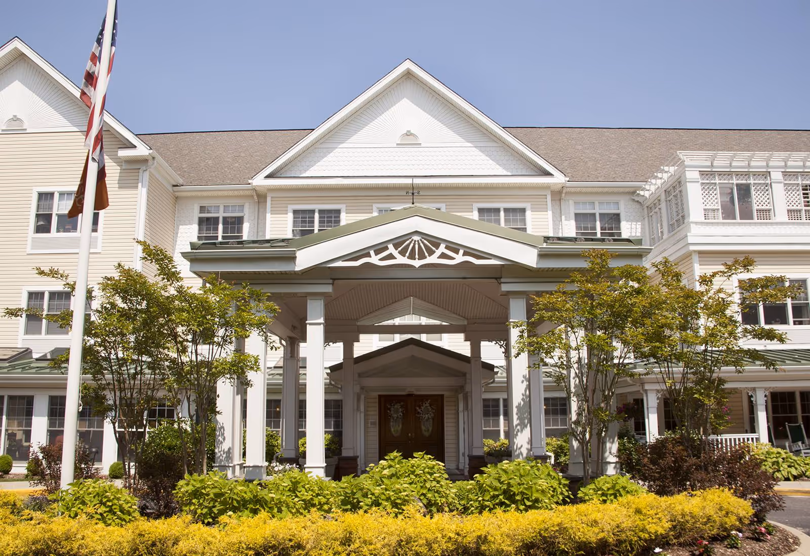 Front exterior view of a large, multi-story senior living facility building with a covered entrance supported by white columns, surrounded by green shrubs and trees under a clear blue sky.