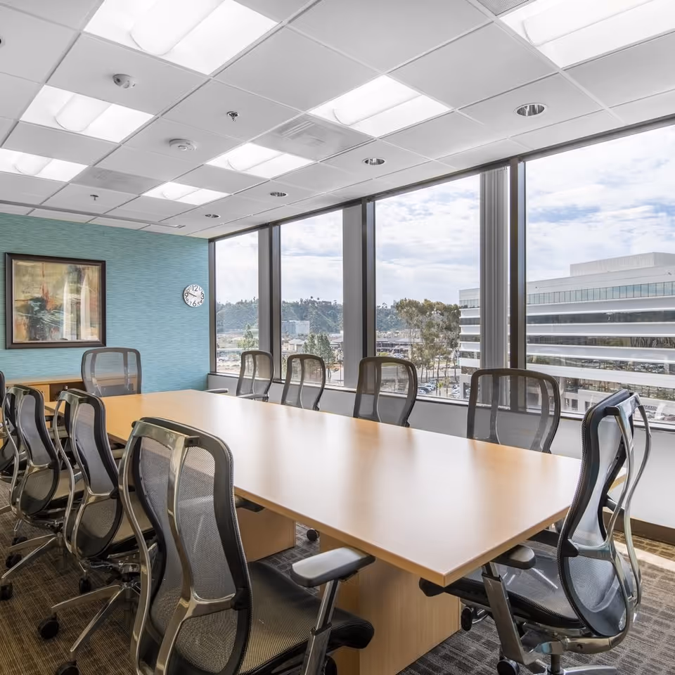 A bright conference room with a long wooden table, mesh chairs, and expansive window views.