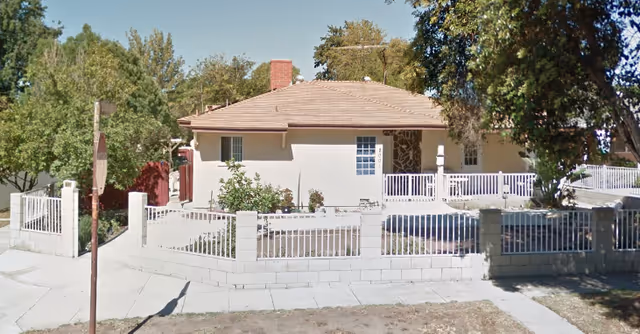 This is a single-story beige home with a white fence, front porch, and surrounding trees.