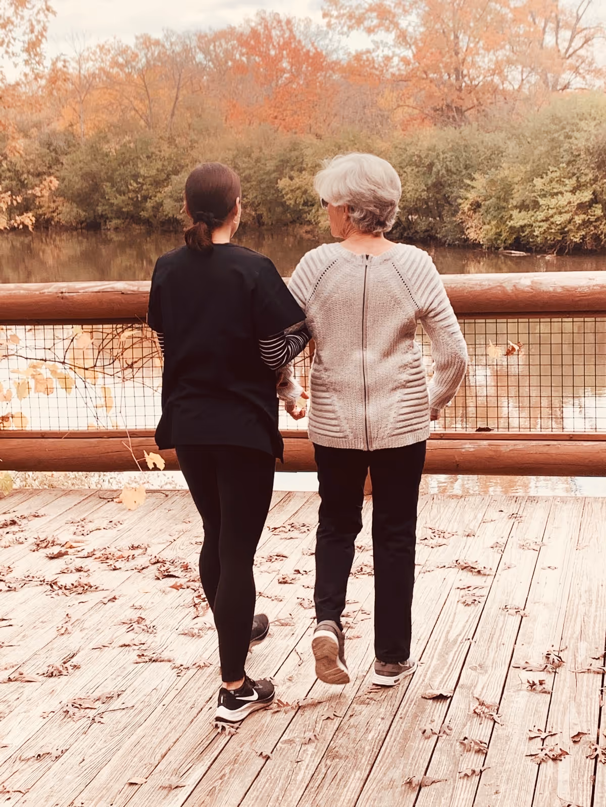 Two people walk on a wooden deck beside autumn trees and water.