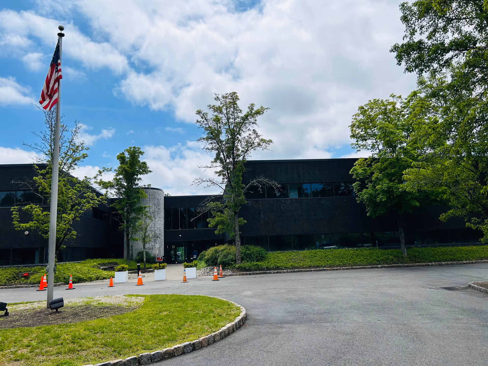 A modern dark building exterior with trees, flagpole, and traffic cones at entrance.