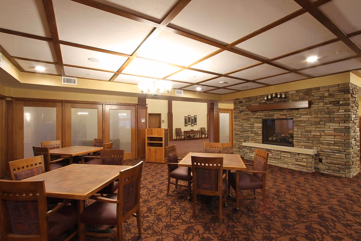 A cozy common area with wooden tables and chairs arranged on a patterned carpet. The room features a stone fireplace on the right wall and a ceiling with wooden beams. There is a chandelier hanging from the ceiling and frosted glass doors along the left wall. In the background, there is a small seating area with two chairs and a table.