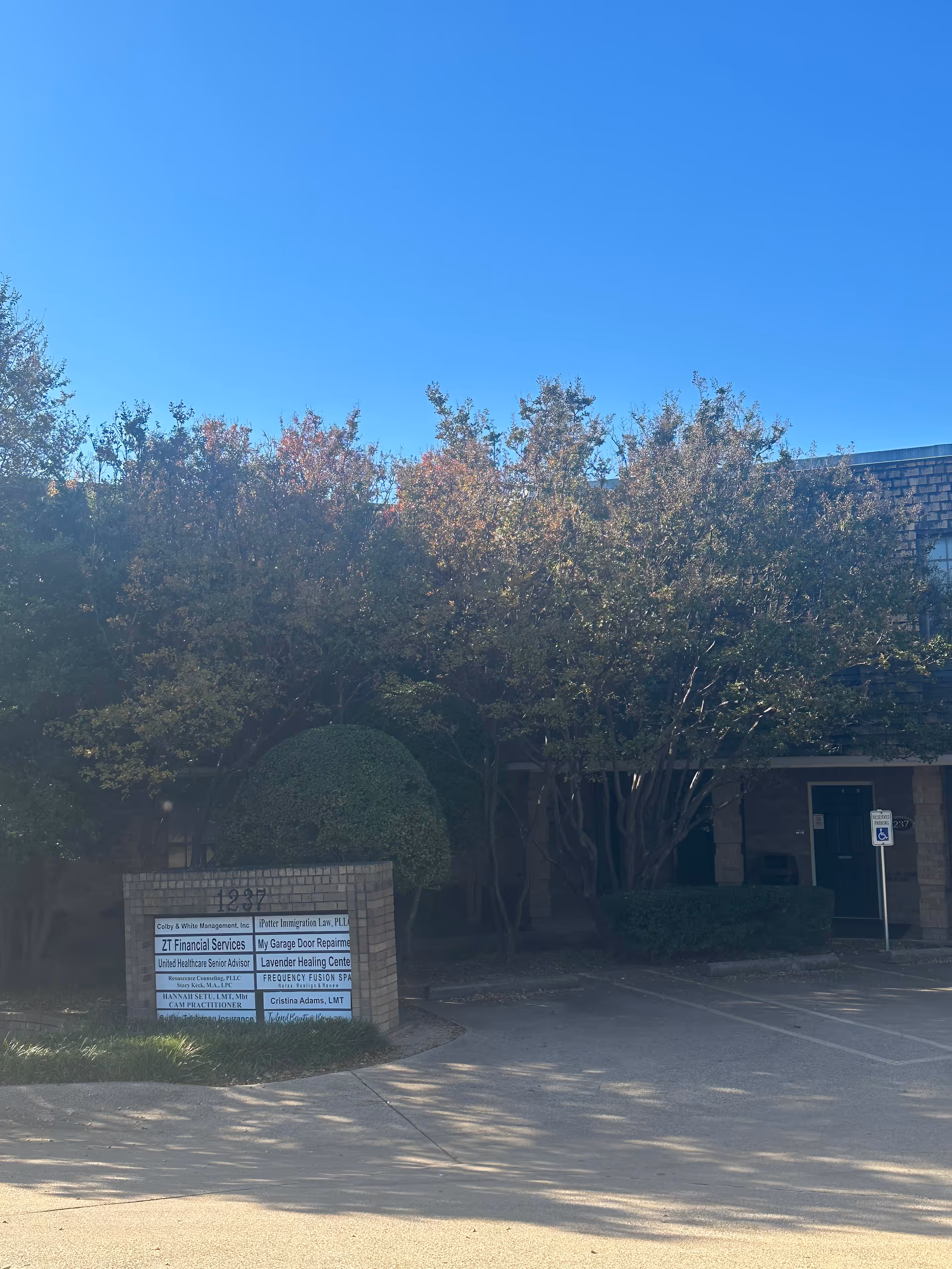 Outside view of a facility entrance with trees, shrubs, and a sign near the driveway.