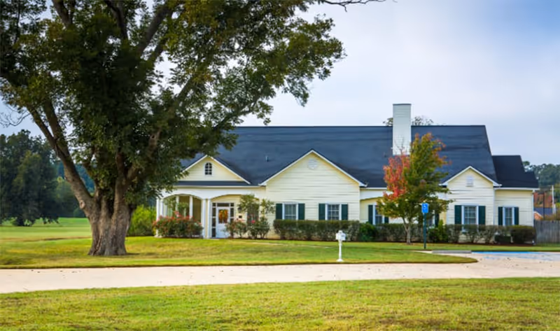 Single-story pale yellow building with a dark roof, front lawn, large tree, and driveway in front.