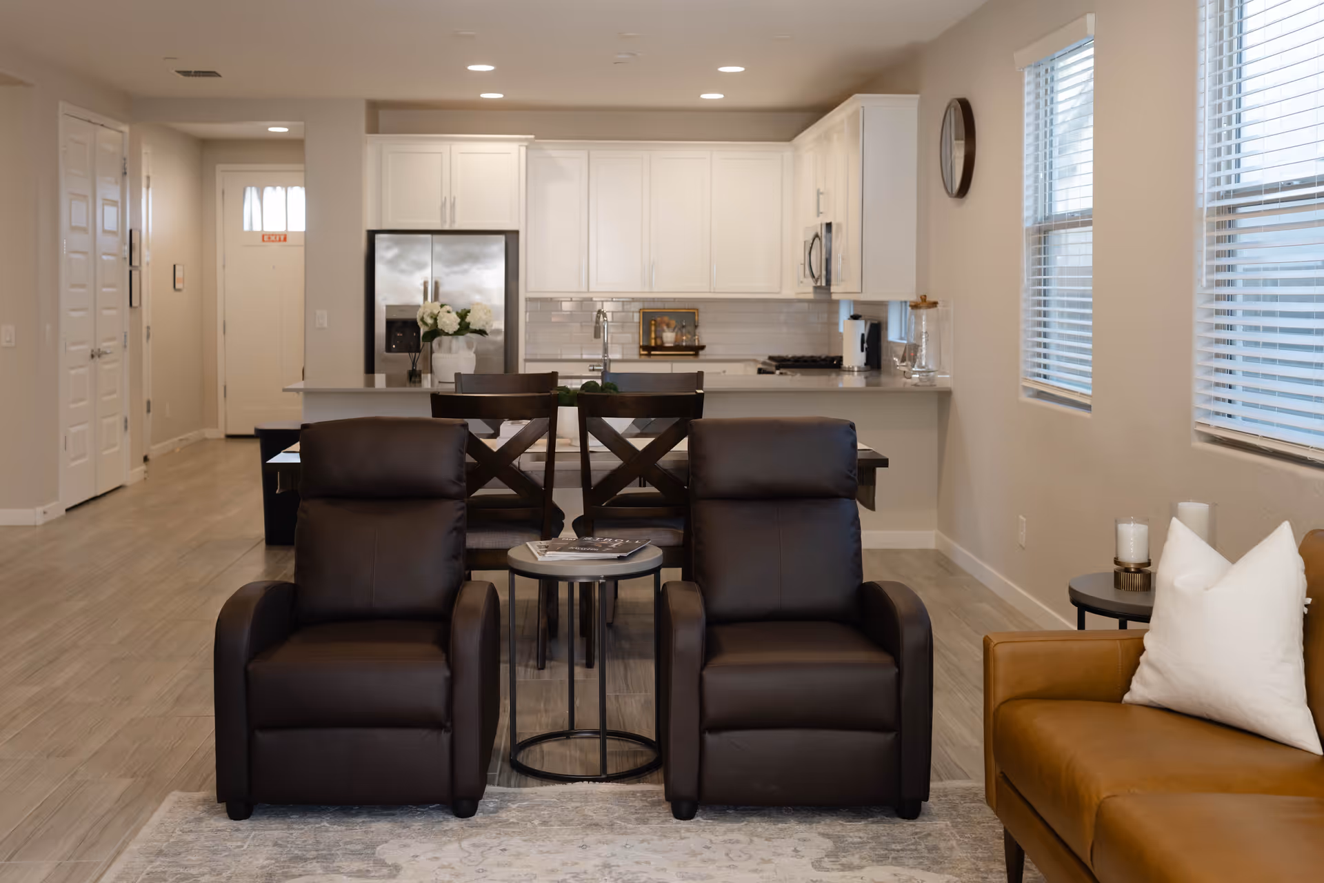 Bright modern living area with two leather recliners and a kitchen in the background.
