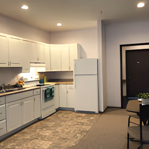 Interior view of a kitchen area with white cabinets, a white refrigerator, a stove with a green towel hanging on the handle, and a countertop with various items. The kitchen floor has a patterned linoleum surface, and the adjacent area is carpeted with a table and chairs visible near a dark doorway.