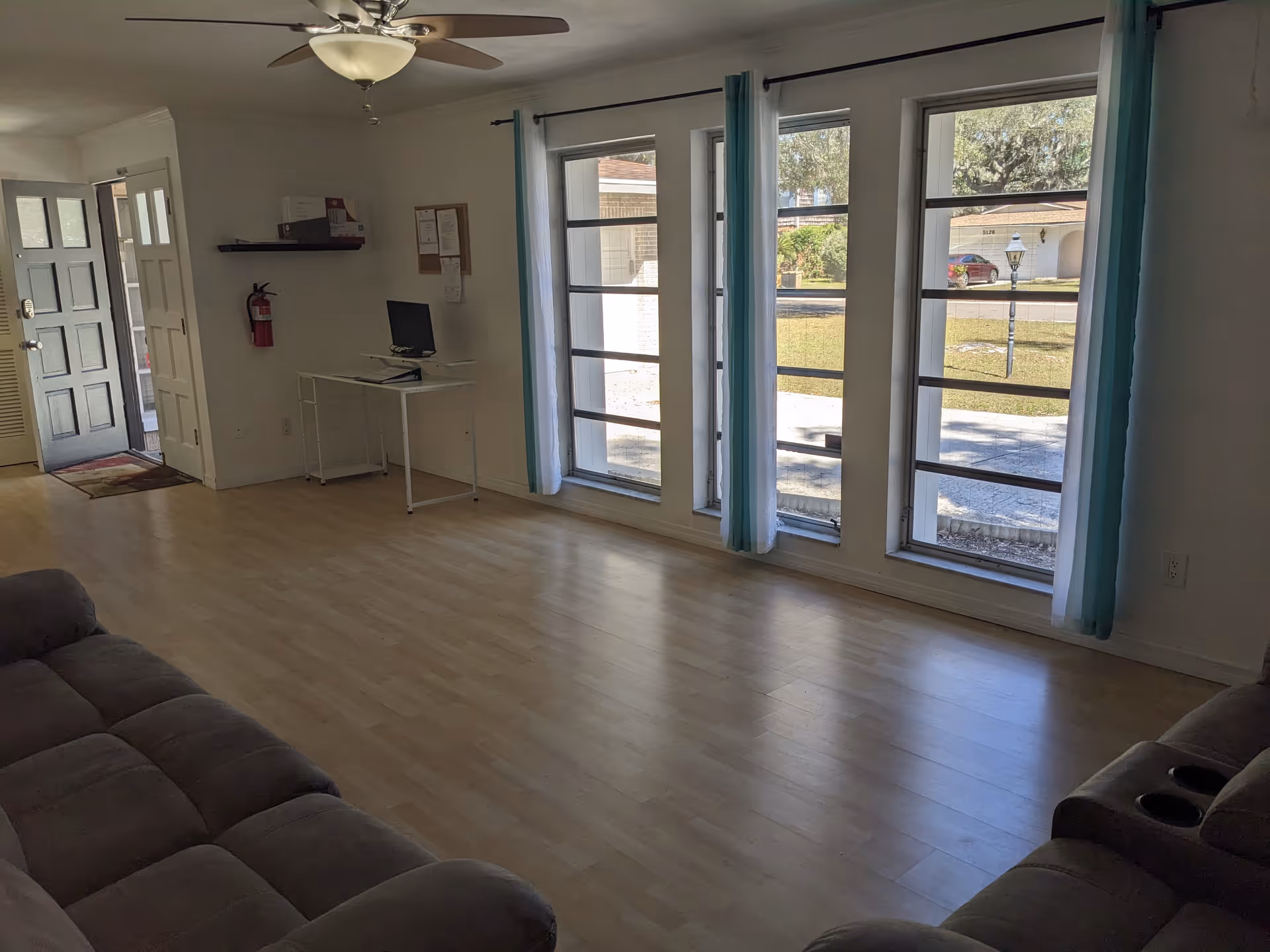 Bright living room with large windows, light wood floors, ceiling fan, and gray sectional seating.