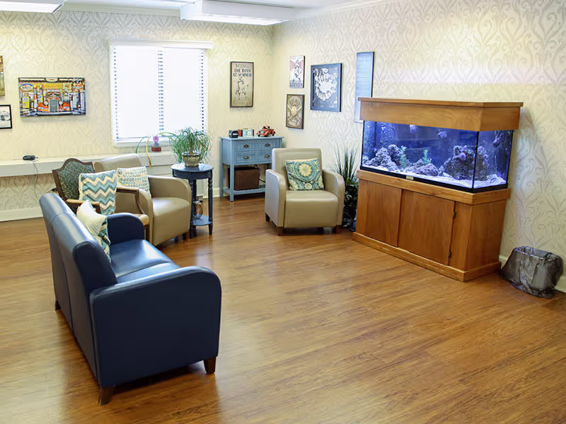 Bright communal lounge with sofas and armchairs, decorative pillows, wall art, plants, and a large aquarium.
